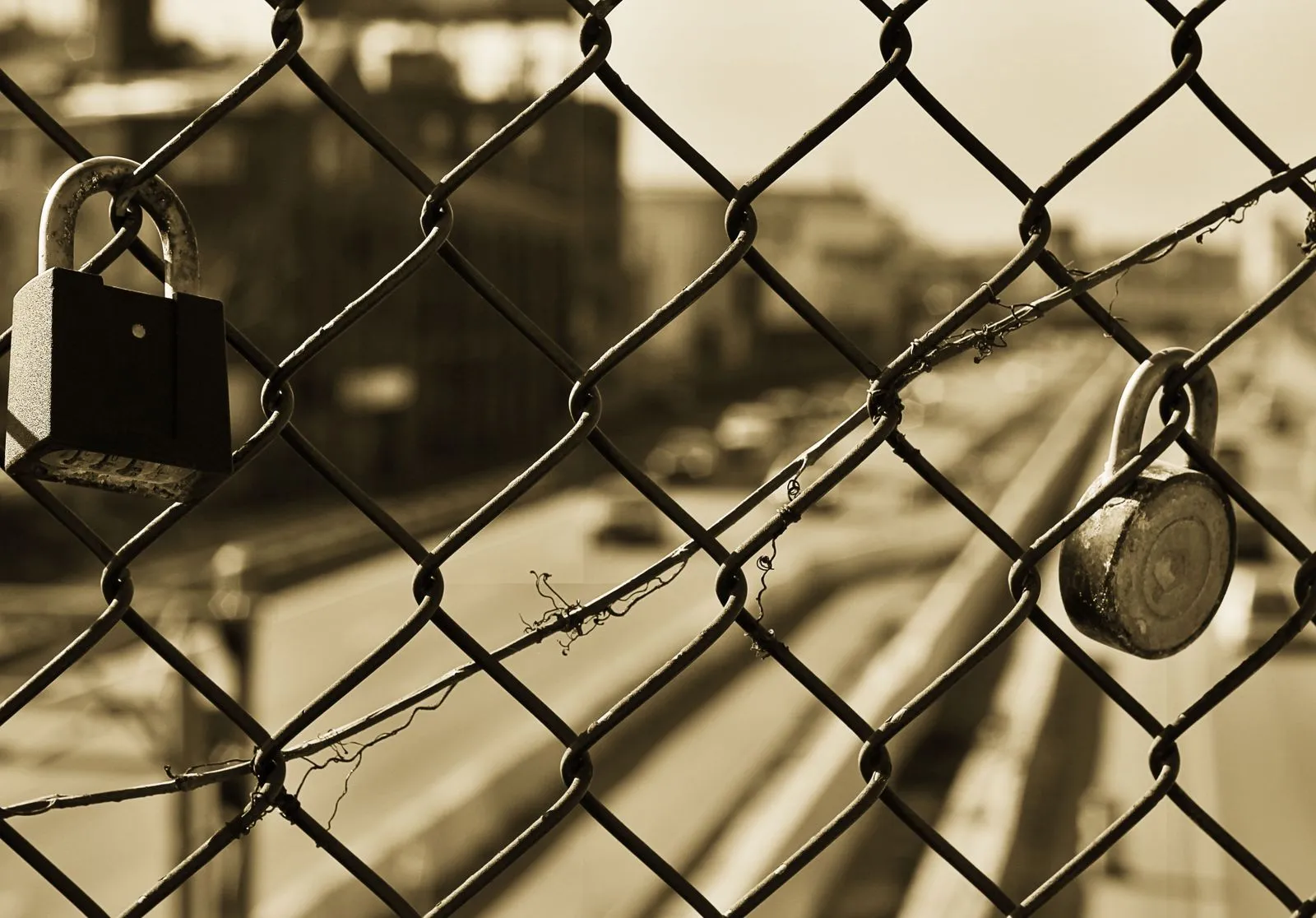 Sepia image of a chain link fence with two locks overlooking a blurred urban street.
